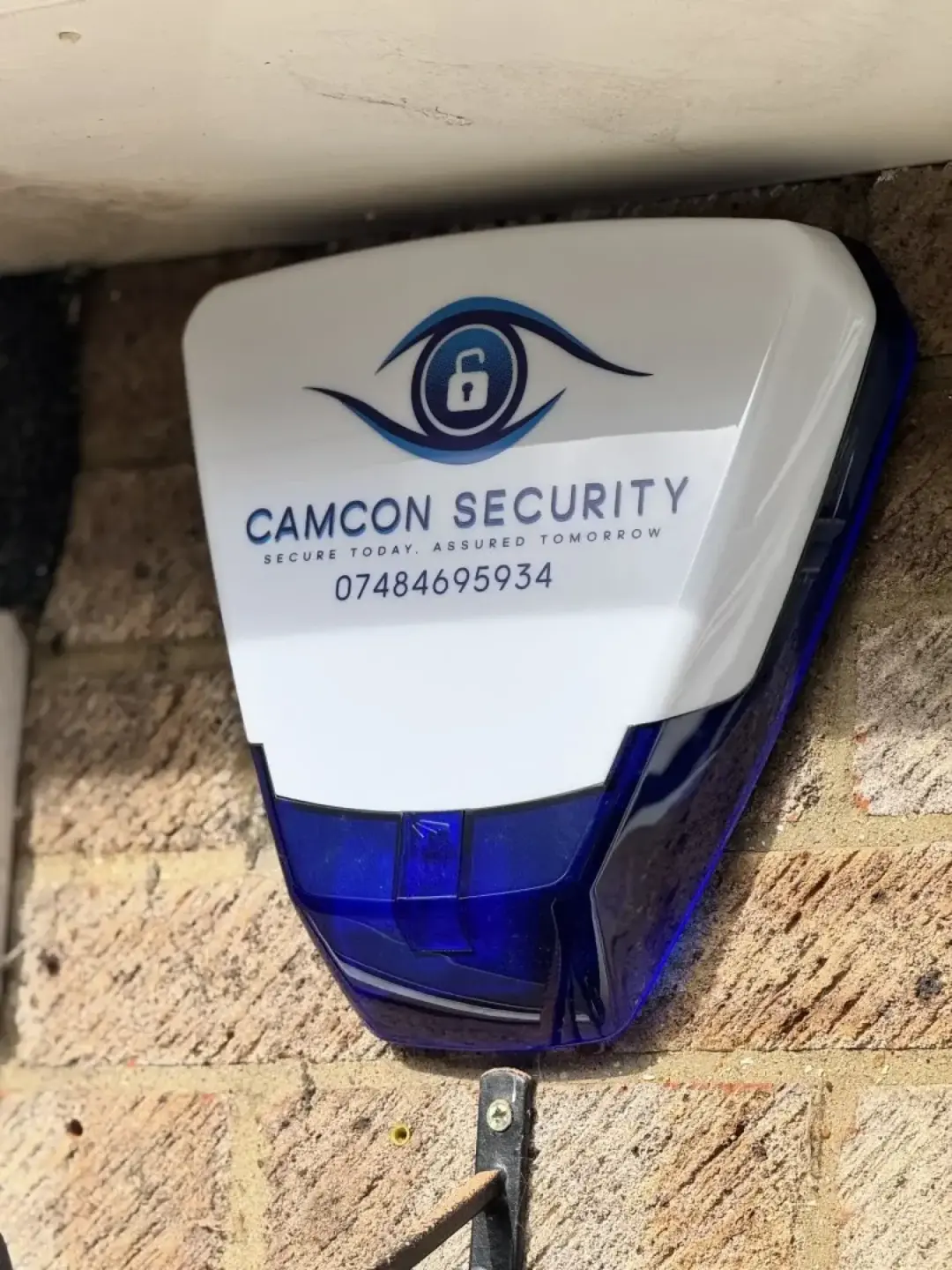 A Camcon Security alarm sounder box fitted under a soffit on a brick wall, with the company phone number on the cover